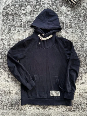 Mörkblå hoodie med snörning - Snygg mörkblå hoodie med grov beige snörning i huvan samt knappar och en liten patch vid fickan. Hoodien har känguruficka framtill och ribbade muddar vid ärmar och nederkant. Perfekt för en avslappnad streetwear-look.