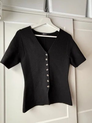 Svart v-ringad topp med knappar - Stilren svart topp från gina tricot i storlek S. Modellen har korta ärmar och en figurnära passform. Den är använd fåtal gånger, säljer då den inte kom till användning💗