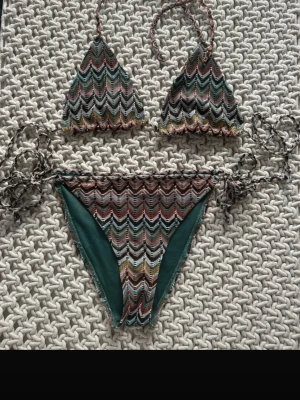 Mönstrad trekantsbikini med knyt - Säljer en snygg trekantsbikini med zigzagmönster i grönt, vitt, gult och rosa. Både överdel och underdel har knytband i sidorna och nacken. Bikinin har en cool retrovibe och är perfekt för stranden eller poolen. Köpt på en marknad i marbella!