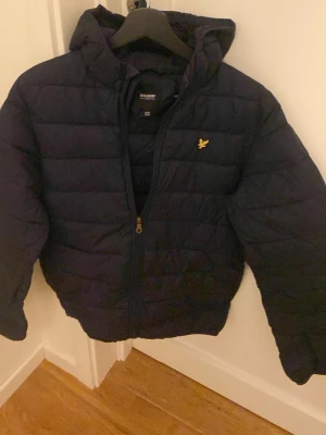 Svart dunjacka från Lyle & Scott - Snygg svart dunjacka från Lyle & Scott Junior med huva och dragkedja. Jackan har det klassiska gula logomärket på bröstet och quiltad design som håller värmen. Perfekt för kalla vinterdagar och riktigt stilren look.