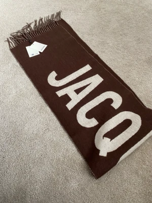 Brun Jacquemus halsduk Virgin Wool - Säljer en brun halsduk från Jacquemus med stor vit logotyp längs hela halsduken. Halsduken har fransar i ändarna och är tillverkad i ett mjukt material som känns lyxigt och trendigt. Perfekt accessoar för att lyfta din outfit.
