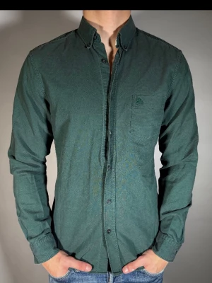 Mörkgrön skjorta med bröstficka - Stilren mörkgrön skjorta med klassisk krage och knappstängning framtill. Skjortan har en bröstficka på vänster sida och långa ärmar med manschetter. Perfekt till jeans för en avslappnad look.