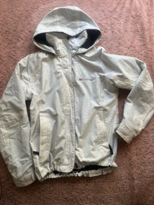  Vindjacka från Everest - Snygg vindjacka från Everest med huva och dragkedja framtill. Jackan har två fickor med dragkedja och justerbar nederkant. Materialet är tunt och vindtätt, perfekt för blåsiga dagar. Diskret logga på bröstet och svart innerfoder i huvan.