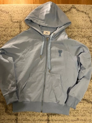 Ljusblå hoodie med dragkedja från Ami - Snygg ljusblå hoodie från Ami med dragkedja framtill och klassisk huva med dragsko. På bröstet finns Ami:s ikoniska hjärtemblem broderat i samma färg. Hoodien har ribbade muddar och fickor framtill. Perfekt för en chill och stilren look helt ny aldrig använd . Storlek S men passar som M