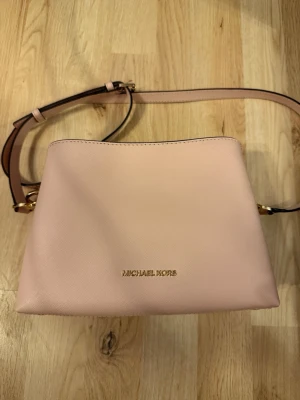 Ljusrosa axelväska Michael Kors - Snygg ljusrosa axelväska från Michael Kors i slätt läder med justerbar axelrem och guldfärgade detaljer. Väskan har en stilren och enkel design med logotypen i guld framtill. Perfekt för dig som gillar minimalistisk och trendig stil. Storlek 22x15.5.