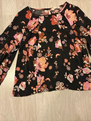 Blommig svart blus från Odd Molly strl 2 (M) - Snygg svart blus från Odd Molly  med färgglatt blommönster i rosa, orange och vitt. Blusen har rund halsringning och långa ärmar med lätt puff. Tillverkad i ett mjukt, tunt tyg som faller fint. Perfekt för dig som gillar färg och mönster!
