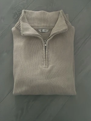 Beige stickad halvzip från C - Snygg beige stickad tröja från Cubus med halvzip och hög krage. Tröjan har ribbade muddar vid ärmslut och nederkant samt en bred ribbad krage. Perfekt för lager på lager och enkel att matcha med olika outfits.