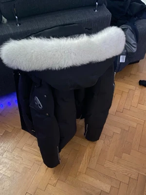 Svart moose knuckle dunjacka med vit päls - Snygg svart dunjacka med stor huva och vit fluffig pälskant. Jackan har flera fickor med dragkedja och tryckknappar samt detaljer i metall. Perfekt för kalla vinterdagar och riktigt varm tack vare tjockt dunfoder.