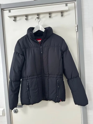 EDC by Esprit jacka - Svart jacka från EDC by Esprit. Jackan har en dragkedja framtill och två fickor med dragkedja i midjehöjd. Kragen är ribbad och insidan av jackan är fodrad med ett mönster av EDC-logotypen. Jackan har även en liten röd etikett med EDC-logotypen längst ner på vänster sida.