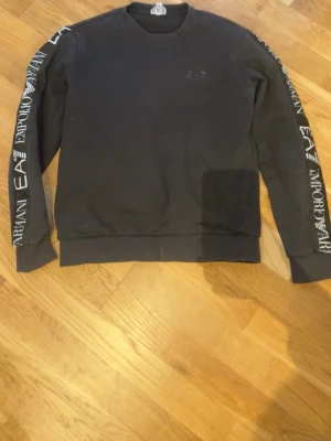 Svart sweatshirt från Emporio Armani - Svart sweatshirt från Emporio Armani EA7 med tryckta loggor längs båda ärmarna. Klassisk rund halsringning och ribbade muddar vid ärmslut och nederkant. Perfekt för en avslappnad och trendig look.