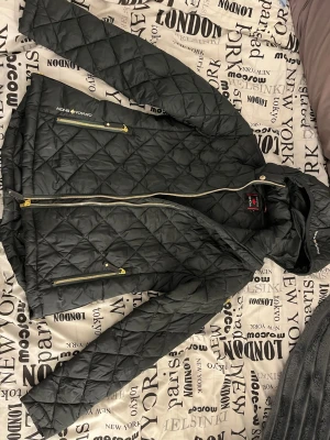 Svart dunjacka från Canada Snow - svart dunjacka från Canada Snow med quiltat mönster och guldfärgade dragkedjor. Jackan har huva och två praktiska fickor framtill. 