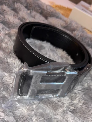 Svart skärp med stor silverfärgad H-spänne - Snyggt svart bälte i skinn med en stor, silverfärgad H-formad spänne. Bältet har vita sömmar längs kanterna som ger en stilren kontrast. Perfekt accessoar för att lyfta vilken outfit som helst.