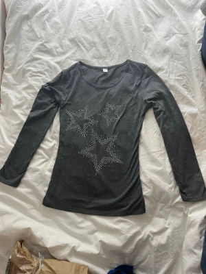 Mörkgrå långärmad topp med stjärnor - Mörkgrå långärmad topp med rund halsringning och tre stora stjärnor framtill dekorerade med små silvriga nitar. Tunn och mjuk kvalitet, figurnära passform. Perfekt för dig som gillar lite extra detaljer på vardagslooken.