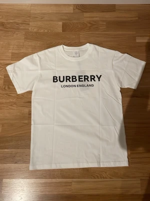 Vit Burberry t-shirt med tryck - Vit t-shirt med svart texttryck 'BURBERRY LONDON ENGLAND' på bröstet. Klassisk rund halsringning och korta ärmar. Tillverkad i mjuk bomull för en clean och stilren look.