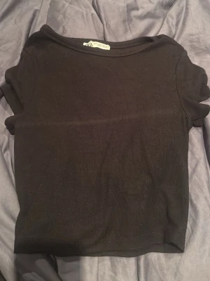 Svart ribbad croppad topp - En svart, croppad topp från Zara med ribbad struktur. Toppen har korta ärmar och en rund halsringning, perfekt för att styla med höga jeans eller kjol. Enkel och stilren design som passar till många olika looks.