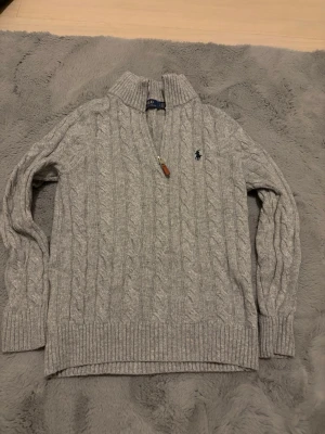 Grå kabelstickad tröja Polo Ralph Lauren - Snygg grå kabelstickad tröja från Polo Ralph Lauren med halv dragkedja och hög krage. Tröjan har ribbade muddar och nederkant samt den klassiska broderade loggan på bröstet. Perfekt för lager på lager. Ny pris ca 2000kr 