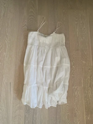 Vit sommarklänning med volanger - Luftig vit kortklänning med tunna axelband och smock upptill. Klänningen har flera lager med volanger och är gjord i ett lätt, skrynkligt bomullstyg som passar perfekt för varma dagar.