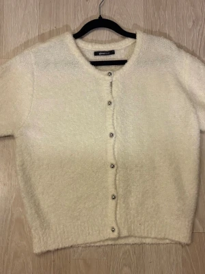 Krämvit kofta från Gina Tricot - Mysig krämvit kofta från Gina Tricot med rund halsringning och silvriga knappar framtill. Koftan är stickad i ett fluffigt material och har kortare ärmar, perfekt för lager-på-lager-stil.