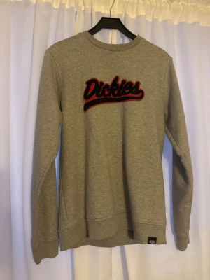 Grå sweatshirt från Dickies - Snygg grå sweatshirt från Dickies med broderad röd och svart logga på bröstet. Tröjan har rund halsringning, ribbade muddar vid ärmslut och nederkant samt en avslappnad passform. Perfekt för en casual streetwear-look.
