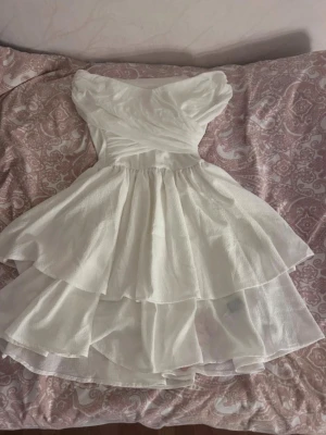 Vit kort volangklänning - Söt vit kortklänning med volanger och offshoulder-design. Klänningen har en omlottliknande överdel och dubbla lager i kjolen för extra volym. Perfekt för sommarens alla tillfällen.