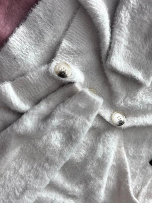 Ljusgrå fluffig kofta med knappar - Supermjuk och fluffig ljusgrå kofta med stora pärlemorknappar. Koftan har en avslappnad passform och är tillverkad i ett lurvigt material som känns riktigt mysigt. Perfekt för lager på lager och chill dagar. ( den sitter som en crop top ) 