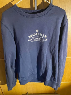 Marinblå sweatshirt från Morris - Snygg marinblå sweatshirt från Morris med trycket 'Morris Gentlemen's Club' på bröstet. Tröjan har rund halsringning, ribbade muddar vid ärmslut och nederkant samt en klassisk, stilren design. Perfekt för en avslappnad men ändå uppklädd look.