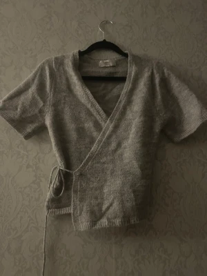 Grå omlottopp från Gina Tricot - Mysig grå omlottopp från Gina Tricot i ullblandning. Toppen har korta ärmar, v-ringning och knyt i sidan för en snygg passform. Perfekt att styla med jeans eller kjol för en trendig look. Kanin finns i hemmet