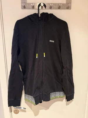 Svart hoodie från BOSS med dragkedja - Svart hoodie från BOSS med dragkedja framtill, huva med snören och små gula detaljer. Nederkanten har ett mönstrat band med BOSS-logga i vitt och gult. Perfekt för en avslappnad och stilren look.