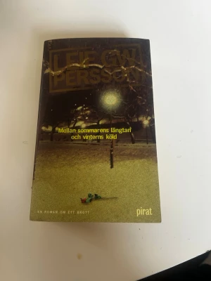 Mellan sommarens längtan och vinterns köld - En kriminalroman av Leif GW Persson med mörkt omslag och gul text. Boken handlar om ett mystiskt dödsfall och polisens utredning i Stockholm. Pocketbok med spännande handling och inslag av samhällskritik.