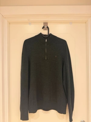 Morris stickad tröja med halvzip - Mörkgrön stickad tröja (100% merinoull) med halvzip och hög krage. Tröjan har en liten broderad logga på bröstet och ribbade muddar vid ärmslut och nederkant. Perfekt för lager på lager och en stilren look.                                                               Modellen är 185cm lång och väger 82kg.