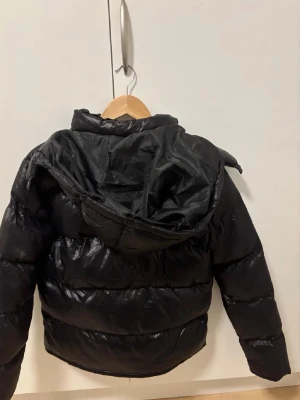 Svart pufferjacka från Polo Ralph Lauren - Snygg svart pufferjacka från Polo Ralph Lauren med huva och dragkedja framtill. Jackan har två fickor med dragkedja och den klassiska röda loggan på bröstet. Tillverkad i glansigt material som ger en modern look. Perfekt för kalla vinterdagar.