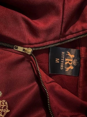 Vinröd hoodie från Rittfors med dragkedja - Snygg vinröd hoodie från Rittfors i storlek M. Hoodien har dragkedja framtill och en stor guldfärgad brodyr på ryggen med lejon, krona och logga. Tillverkad i ett glansigt, mjukt material som känns lyxigt. Perfekt för dig som vill sticka ut med stil.