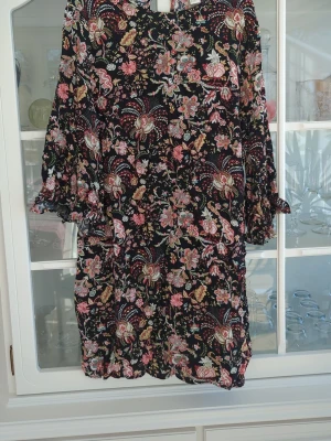 Blommig svart kortklänning - Svart kortklänning med färgglatt blommönster i rosa, rött, grönt och beige. Klänningen har trekvartsärm med volangdetaljer och en lätt v-ringning. Perfekt för dig som gillar boho-vibbar och vill ha en luftig och snygg look.