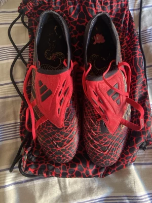 Adidas Predator fotbollsskor röd/svart - Säljer ett par Adidas Predator fotbollsskor i svart och rött med coolt reptilmönster och röda snören. Skorna har låg profil, klassisk Predator-design och slitstark syntetisk ovandel. Perfekta för dig som vill sticka ut på planen.