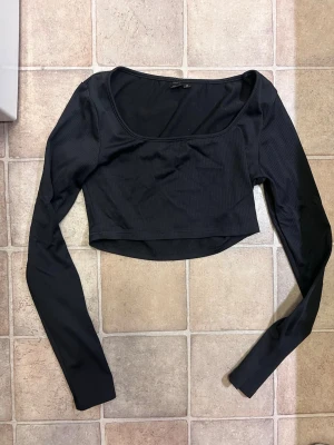 Svart croppad långärmad topp - Trendig svart croppad topp med långa ärmar och rundad urringning. Tillverkad i ribbad stretchig bomull som sitter snyggt och bekvämt. Perfekt att styla med höga jeans eller kjol för en cool look.