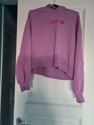 Lila cropped hoodie med tryck - Säljer en lila cropped hoodie med huva och trycket 'JULIA' samt ett rosa hjärta på bröstet. Hoodien har långa ärmar med muddar och en avslappnad passform. Perfekt för dig som gillar en chill och trendig stil. Har en liten fläck på ena armen.