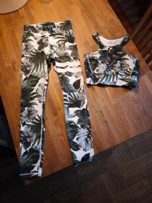 Träningsset med tropiskt mönster Stronger - Snyggt träningsset från Stronger med tropiskt bladmönster i grönt, vitt och svart. Setet består av ett par tights med hög midja och en matchande sport-bh med breda axelband. Perfekt för gymmet eller yoga. Materialet är stretchigt och följsamt.