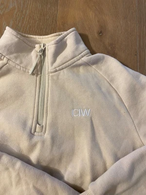 Beige halfzip hoodie från ICIW - Snygg beige hoodie från ICIW med halv dragkedja framtill och broderad logga på bröstet. Tillverkad i mjukt sweatshirtmaterial och har en hög krage. Perfekt för en avslappnad och sportig stil.