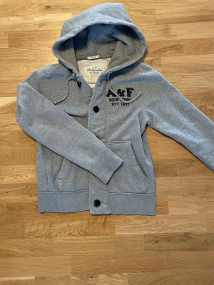 Grå hoodie från Abercrombie & Fitch - Snygg grå hoodie från Abercrombie & Fitch med broderad A&F New York-logga på bröstet och knäppning framtill. Tröjan har huva med dragsko, ribbade muddar och två fickor. Tillverkad i mjukt bomullsmaterial för en avslappnad look.