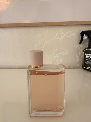 Burberry Her Eau de Parfum 100ml - Burberry Her Eau de Parfum i en stilren, rektangulär glasflaska med ljusrosa lock och gulddetalj. Doften har en mjukt rosa färg och flaskan har Burberry-loggan ingraverad längst ner. Perfekt för dig som gillar eleganta och moderna parfymer.