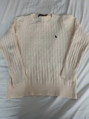 Vit kabelstickad tröja från Ralph Lauren - Kabelstickad tröja i mjuk vit ullblandning från Ralph Lauren. Klassisk rund halsringning, långa ärmar och ribbade muddar. Ikoniska lilla Polo-loggan broderad på bröstet. Perfekt för lager på lager och en clean look. Är som storlek 164.