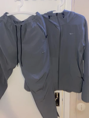 Grått träningsset från Nike - Snyggt grått träningsset från Nike med matchande jacka och byxor. Jackan har huva, dragkedja och liten Nike-logga på bröstet. Byxorna har resår i midjan, svarta snören och Nike-logga på benet. Perfekt för löpning eller gym. jacka storlek S byxor storlek M