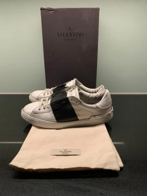 Valentino opens - Snygga vita sneakers från Valentino Garavani med en bred svart läderdetalj över sidan. De har gått lås lite på framsidan av skon! Klassisk låg modell med rund tå, vita skosnören och diskret logga på tungan. Skorna är tillverkade i läder och har en stilren design som sticker ut. Storlek 42,5 men passar storlek 42-43. Medföljer box och 2 dustbags!