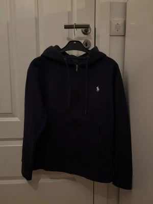 Mörkblå hoodie från Polo Ralph Lauren - Snygg mörkblå hoodie från Polo Ralph Lauren med dragkedja framtill och klassisk vit logga broderad på bröstet. Hoodien har justerbar huva med snören och två fickor framtill. Perfekt för en avslappnad och stilren look.