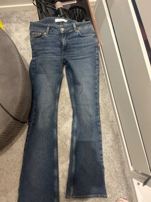 Blå bootcut jeans från Junkyard - Snygga blå bootcut jeans från Junkyard med klassisk femficksmodell och ljusare slitningar framtill. Jeansen har kontrastsömmar och en läderpatch bak i midjan. Perfekta för en avslappnad och trendig look.