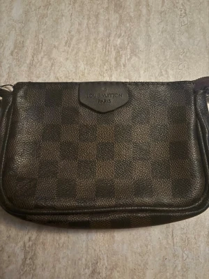 Brun axelväska - Säljer en klassisk brun axelväska med det ikoniska rutmönstret Damier. Väskan har en rektangulär form och är tillverkad i tåligt canvasmaterial med läderdetaljer. Perfekt för dig som gillar lyxiga accessoarer och tidlös design. Saknar kedjan.