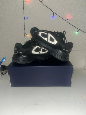 Svarta Dior sneakers med CD-logga - Snygga svarta Dior sneakers med stor vit CD-logga på sidan. Skorna har en chunky sula, mesh och läderdetaljer samt svarta skosnören. Perfekta för dig som vill ha en exklusiv och trendig look. Klassisk low-top modell med tydlig branding.