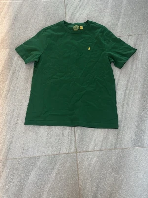 Str-14y. Grön t-shirt från Ralph Lauren - Mörkgrön t-shirt från Ralph Lauren med klassisk passform och korta ärmar. Liten gul broderad logga på bröstet. Tillverkad i mjuk bomull som känns skön mot huden. Perfekt till jeans eller shorts för en clean och enkel stil. Aldrig använt den!