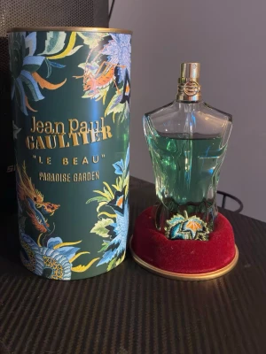 Jean Paul Gaultier Le Beau Paradise Garden - Unik parfymflaska formad som en manlig torso i glas med turkosgrön vätska och guldfärgad kork. Kommer i en dekorativ grön tub med tropiskt mönster och texten 'Le Beau Paradise Garden'. Flaskan står på en röd sammetsliknande bas med broderad detalj. Sällan använd.