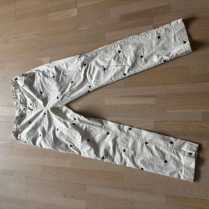 Vita byxor med svart mönster Morris - Unika vita bomulls byxor från Morris med små svarta broderade detaljer över hela tyget. Byxorna har rak passform och elastisk midja för extra komfort. Perfekta för dig som vill sticka ut med en stilren men ändå lekfull look.